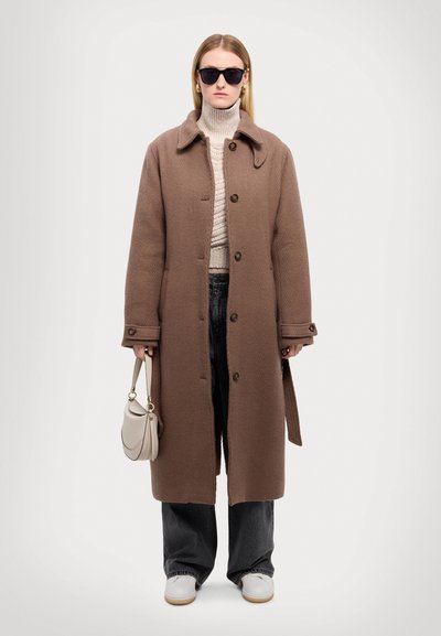 Bruine visgraatjas met een kraag, knoopsluiting, crèmekleurige trui, zwarte broek en witte sneakers; met een lichtbeige handtas.