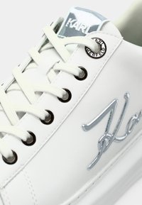 SNEAKER de couro branco com detalhes em logo prateado, ilhós redondos pretos e atacadores brancos planos. Textura suave com um design minimalista.