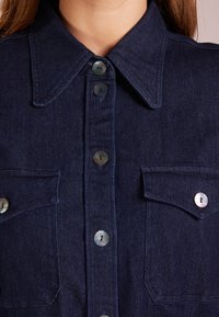 Tumma denim-paita, jossa on terävä kaulus, nappikiinnitys edessä, kaksi rintataskua ja helmenkaltaiset, sileäpintaiset napit.