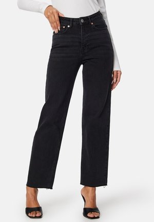 Jeans en denim noir taille haute avec une coupe droite, présentant un design classique à cinq poches et des détails de couture subtils, portés avec des talons ouverts.