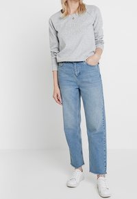 Sudadera gris clara con logo en relieve, combinada con jeans de mezclilla azul claro y de pierna recta de cintura alta. Zapatillas blancas completan el look.