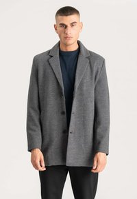 Ung mand med kort hår iført en grå uldfrakke med knapper over en mørk crew-neck sweater og sorte bukser, stående foran en ensfarvet baggrund.
