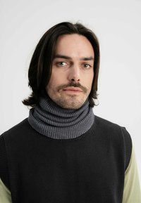 Grijze geribbelde coltrui met hoge kraag, gelaagd onder een zwart mouwloos topje. Model heeft donker haar en een lichte huid.