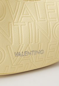Geltona odinė rankinė su iškilmiu užrašu "VALENTINO" ir sidabro spalvos detalėmis, pasižyminti lygaus paviršiaus ir struktūrine forma.