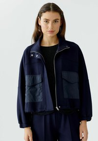 Marineblauwe cropped jas met een hoge kraag, ritssluiting aan de voorkant en elastische manchetten. Twee grote opgestikte zakken aan de voorkant met een gladde textuur.