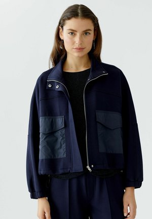 Marineblauwe cropped jas met een hoge kraag, ritssluiting aan de voorkant en elastische manchetten. Twee grote opgestikte zakken aan de voorkant met een gladde textuur.