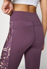 Vrouw past paarse high-waist leggings aan met een bruin en wit abstract zijpatroon, in combinatie met een wit cropped top, tegen een effen achtergrond.