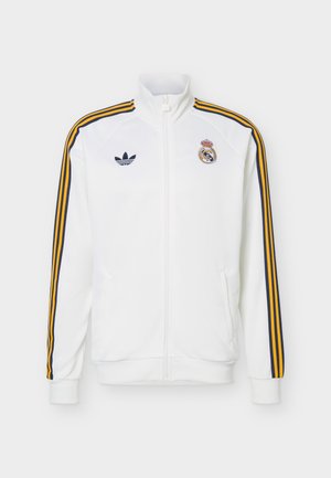 Witte jas met rits met oranje en zwarte strepen op de mouwen, Adidas-logo op de linkerborst en Real Madrid-wapen op de rechterborst.