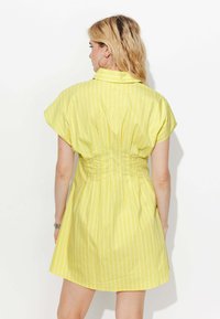 Femme blonde avec des boucles d'oreilles créoles portant une robe jaune rayée à manches courtes, se tenant face à un mur blanc, le dos à l'appareil photo.