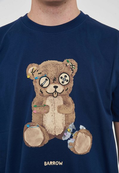 T-shirt blu navy con una grafica di un orsetto in peluche, con spille da balia e dettagli cuciti. Testo "BARROW" stampato sotto l'orsetto. Tessuto morbido.