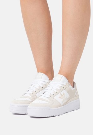 Piernas con zapatillas Adidas blancas y beige con cordones, sobre un fondo claro y liso.