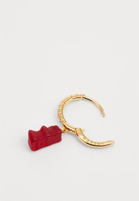 Boucle d'oreille en métal doré avec un charme en forme de ours en gélatine rouge portant l'inscription "HARIBO" et un design texturé sur le hoop.