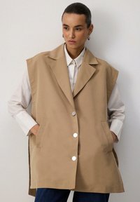 Beige ilma varrukat vest, millel on kohandatud disain, suured kraed, hõbedased nupud ja küljetaskud, mille peal on valge särk.