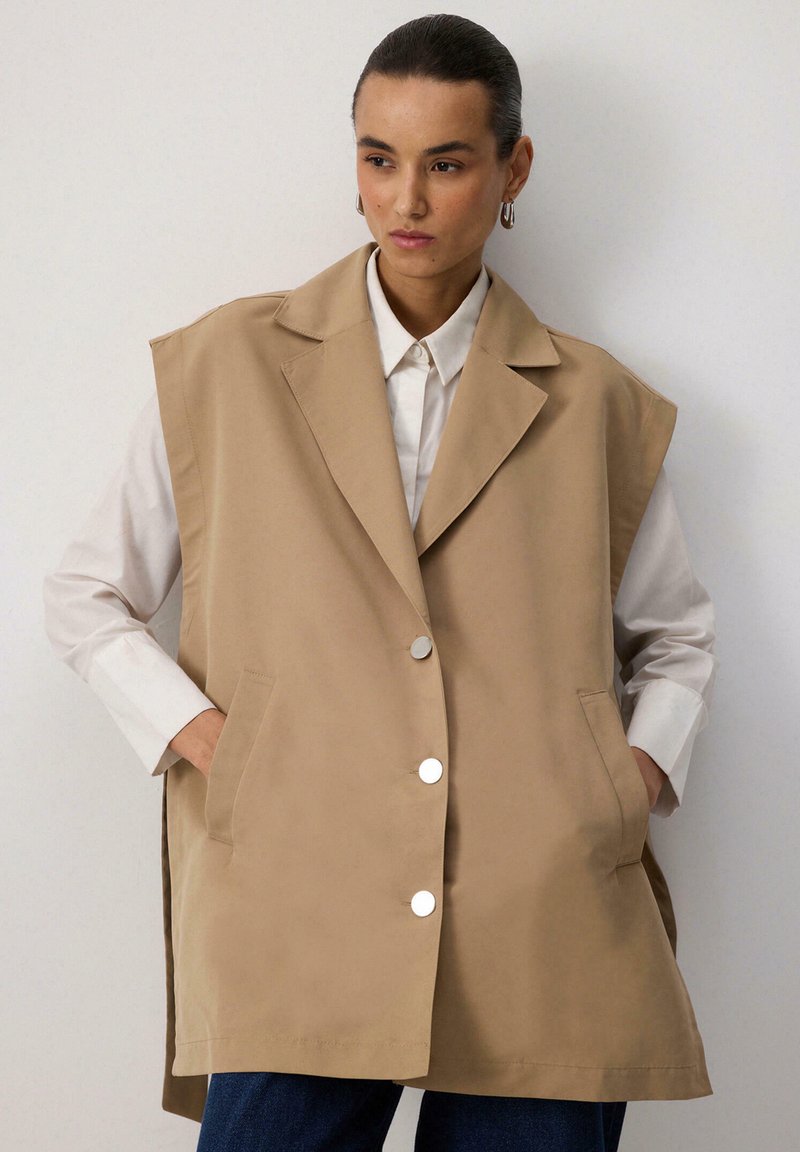 Beige ilma varrukat vest, millel on kohandatud disain, suured kraed, hõbedased nupud ja küljetaskud, mille peal on valge särk.