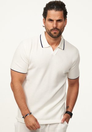 Weißes kurzärmliges Poloshirt aus strukturiertem Stoff, mit einem navyfarbenen Kragen und Akzenten an den Ärmeln, ergänzt durch ein Armband am Handgelenk.