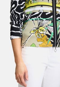 Zwart-wit gestreepte lange mouwen top met een bloemenpatroon in geel en groen, met een ritssluiting aan de voorkant en ribbeltextuur. Gecombineerd met witte broek.