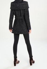 Abrigo de lana de mezclilla negro, de corte grande, con un amplio cuello y detalle de cinturón, combinado con leggings negros y botines marrones. Acabado texturizado.