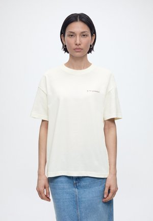 Femme aux cheveux courts et foncés, vêtue d'un t-shirt blanc à manches courtes et d'une jupe en denim bleu clair, debout devant un fond blanc uni.