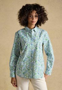 Vrouw met krullend haar en bril, gekleed in een blouse met lange mouwen, met een bloemenpatroon in blauw, groen en wit, gecombineerd met lichtgekleurde broek.