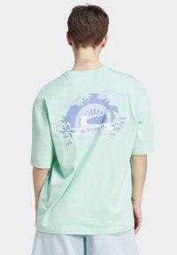 Mintgrön oversized t-shirt med korta ärmar. På baksidan finns en grafik med palmer och blommönster, samt en Adidas-logotyp i mitten.
