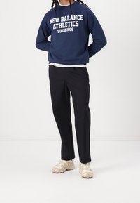 Navy huppari, jossa on valkoiset tekstit "New Balance Athletics", yhdistettynä mustiin housuihin ja vaaleanbeigeihin lenkkareihin.