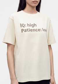Personne portant un t-shirt beige avec un texte marron « "IQ: high Patience: low" » centré sur la poitrine sur un fond uni.