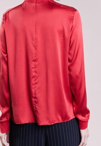 Rote Satinbluse mit langen Ärmeln, hohem Kragen und glatter Textur. Verfügt über eine zentrale Naht im Rücken und einen leicht gerundeten Saum.