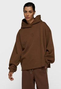 Mujer con sudadera holgada de capucha marrón y pantalones marrones a juego con cordones, de pie contra un fondo liso y claro.