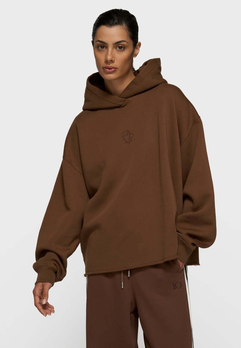 Mujer con sudadera holgada de capucha marrón y pantalones marrones a juego con cordones, de pie contra un fondo liso y claro.