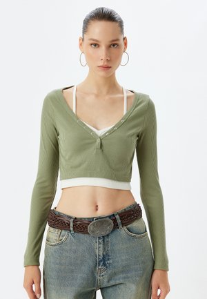 Olivgrünes, geripptes Crop-Top mit langen Ärmeln, das eine V-Ausschnitt und Knopf-Akzente aufweist. Über einem weißen, schichtigen Tanktop getragen, kombiniert mit blauen Jeans.