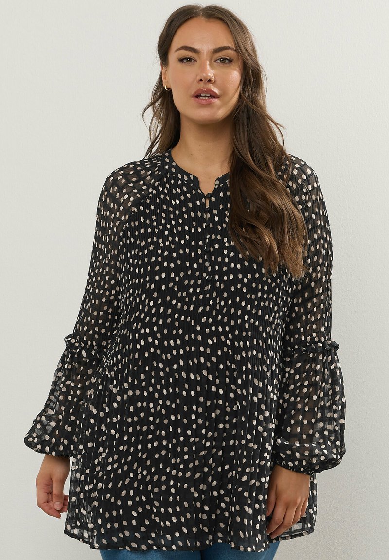 Svart genomskinlig blus med vita prickar, plisserad design, rund halsringning och trumpetärmar. Texturerad tyg ger djup till looken.