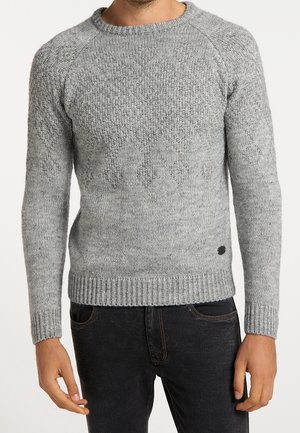 Mann trägt hellgrauen, strukturierten Strickpullover mit Rautenmuster, kombiniert mit dunkler Jeans, vor hellem einfarbigem Hintergrund.