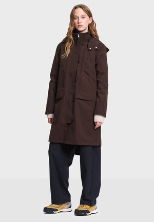 Femme portant un long manteau marron foncé avec de grandes poches, un pantalon large bleu marine, des baskets jaunes et un pull crème, debout face à l'avant.