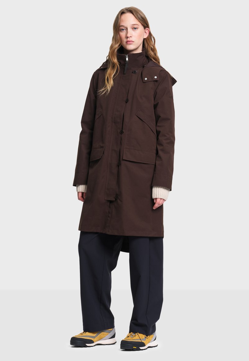 Femme portant un long manteau marron foncé avec de grandes poches, un pantalon large bleu marine, des baskets jaunes et un pull crème, debout face à l'avant.