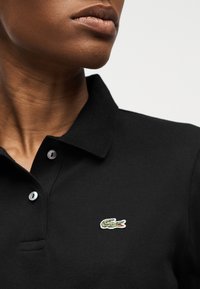 Gros plan d'une personne portant une chemise noire à col avec un petit logo crocodile vert brodé sur la poitrine.