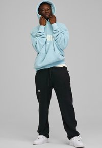 Lys blå hettegenser med hvit tekst, svarte joggebukser med logo, og hvite sneakers. Hette-design med lomme, avslappet passform og snor i livet.