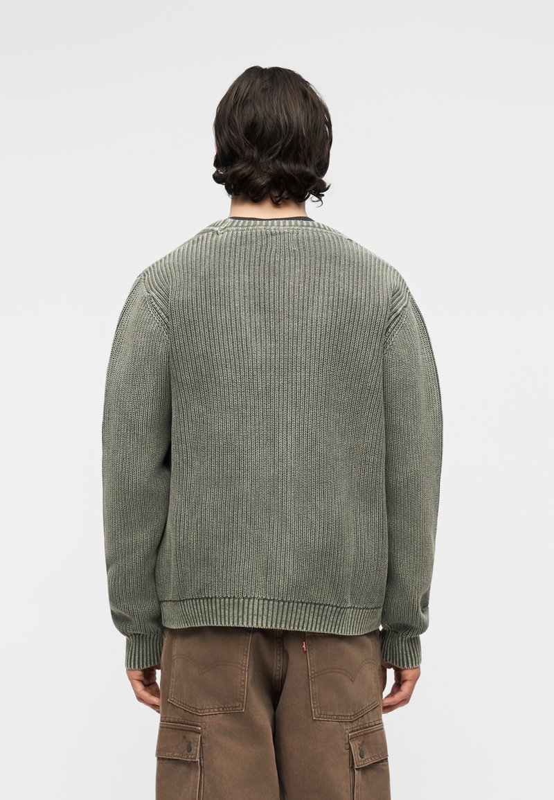 Person mit dunklen Haaren, die einen locker sitzenden grauen Strickpullover und braune Cargohosen trägt, von hinten vor einem einfarbigen Hintergrund gesehen.