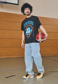 Sort t-shirt med blå og orange grafik, lyseblå jeans og sorte/gule sneakers; holder en rød basketball i en gymsal.