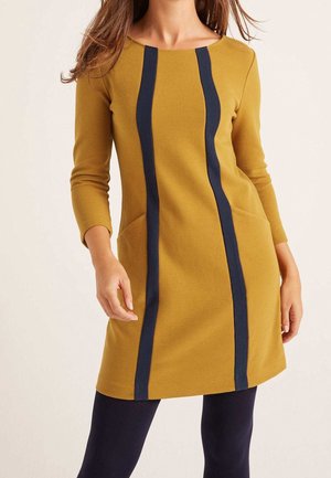 Kobieta ubrana w musztardowożółtą sukienkę z długim rękawem, z dwoma pionowymi granatowymi paskami i kieszeniami po bokach, w połączeniu z czarnymi legginsami.