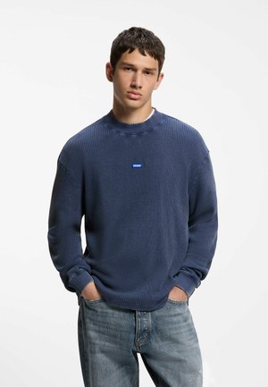 Joven con cabello corto y oscuro lleva un suéter azul texturizado con pequeño logo en el pecho y vaqueros azul claro, manos en los bolsillos, fondo neutro.