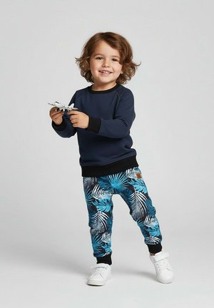 Uśmiechnięte dziecko w granatowej bluzie i niebieskich spodniach w tropikalny wzór trzyma zabawkowy samolot, stojąc na jednolitym szarym tle.