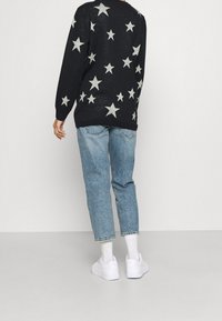 Schwarzer Pullover mit silbernem Sternenmuster, lockere Passform, getragen über hellblauen Jeans, kombiniert mit weißen Turnschuhen und knöchellangen Socken.