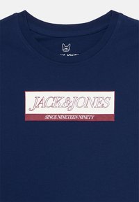 Camiseta de cuello redondo azul marino con un logotipo rectangular blanco y rojo que dice "JACK & JONES" y "DESDE MIL NOVECIENTOS NOVENTA."