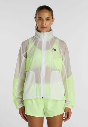 Vrouw die neongele sportshorts draagt en een doorschijnend wit en neongeel jack met rits, staand tegen een effen achtergrond.