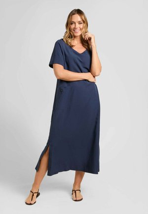 Donna sorridente in piedi che indossa un abito lungo blu navy a maniche corte con spacco laterale e sandali bassi marroni su uno sfondo chiaro uniforme.