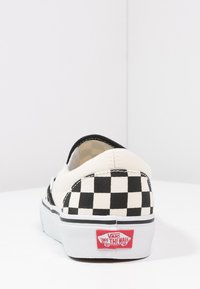 Lærret slip-on sko med et svart og hvitt rutemønster, hvit såle, og rødt Vans-logo på hælen. Enkel, lavprofil design.