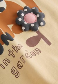 Flores de tela negra con lunares blancos y un centro rosa sobre tela beige con texto marrón que dice ""in the garden"" y un gráfico de planta.