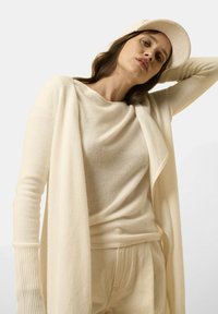 Cárdigan largo de punto suave en color crema, con puños acanalados, superpuesto a un suéter ligero y pantalones a juego. Una gorra de ajuste slim completa el look.