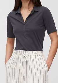 Kvinna i en mörkgrå kortärmad polo skjorta instoppad i vita högmidjade byxor med vertikala svarta ränder och ett knutet midjebälte.