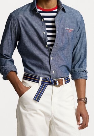 Homme portant une chemise bleue à boutons sur un t-shirt rayé noir et blanc, pantalon blanc avec ceinture rayée bleue, et montre-bracelet marron.